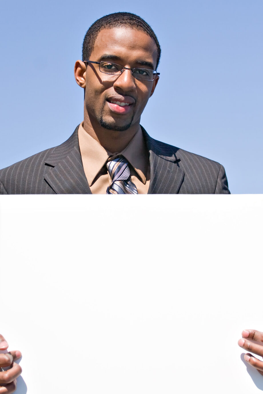 Man in a pinstripe suit and glasses holding a blank white sign against a clear blue sky.