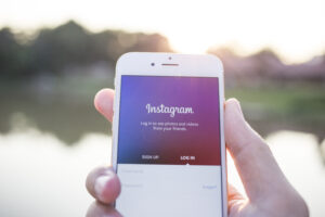 A person holds a smartphone displaying the Instagram login screen with options to "Sign up" and "Log in" against a blurred outdoor backdrop.