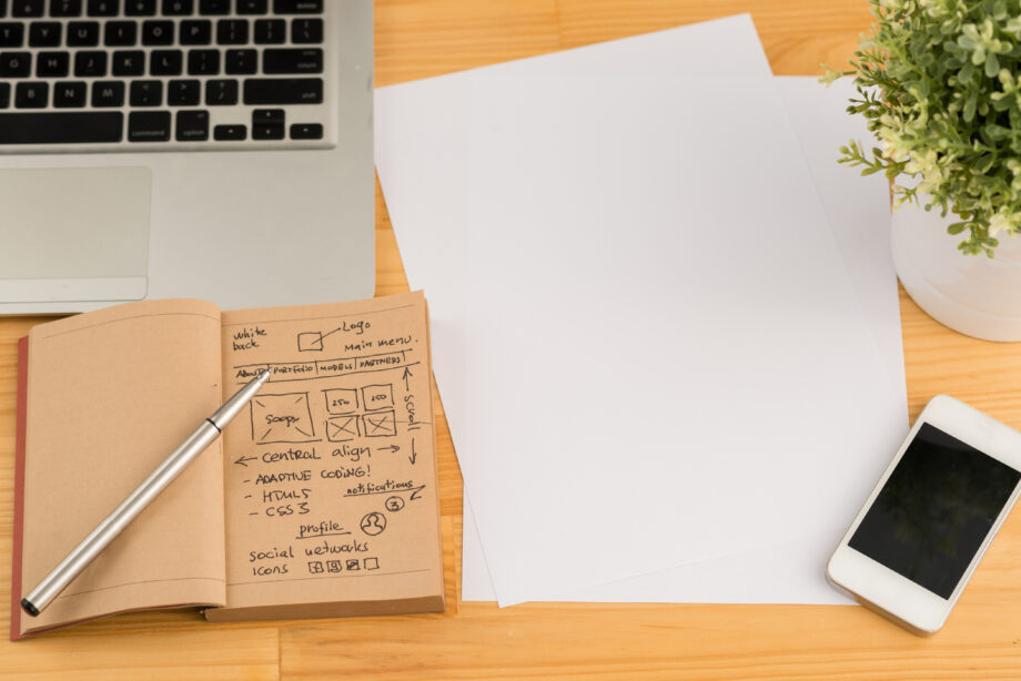 A notebook with web design sketches, a pen, blank papers, a laptop, a smartphone, and a potted plant on a wooden desk.