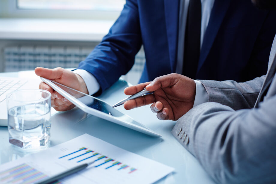 Two people in suits are discussing information displayed on a tablet. A pen, a glass of water, and documents with graphs are on the table.