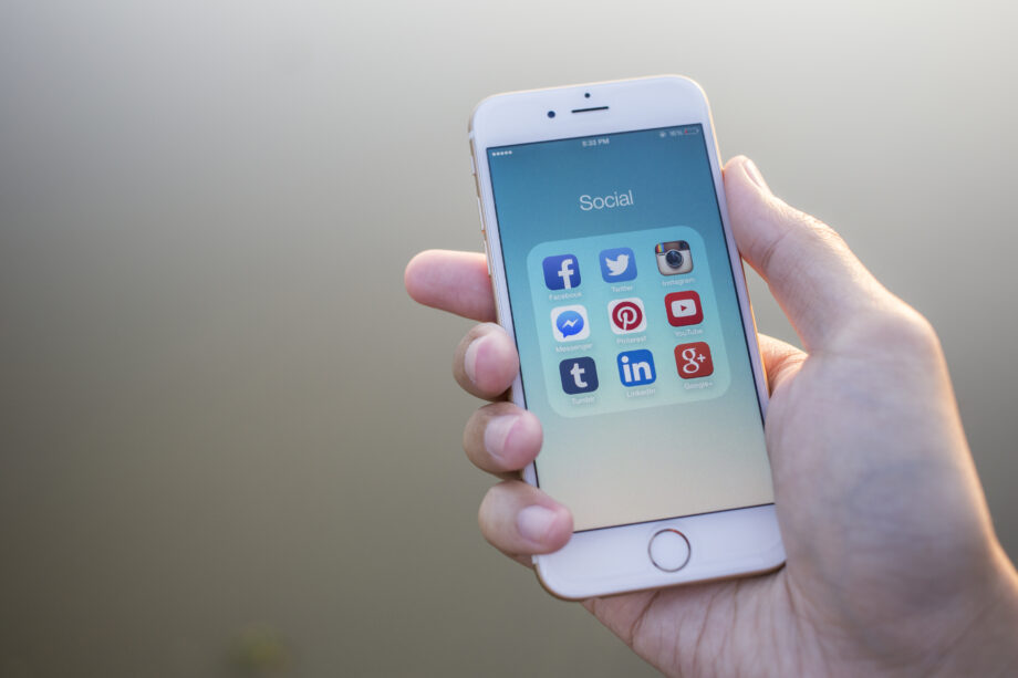 A hand holding a smartphone displaying a folder titled "Social" with various social media app icons on the screen.