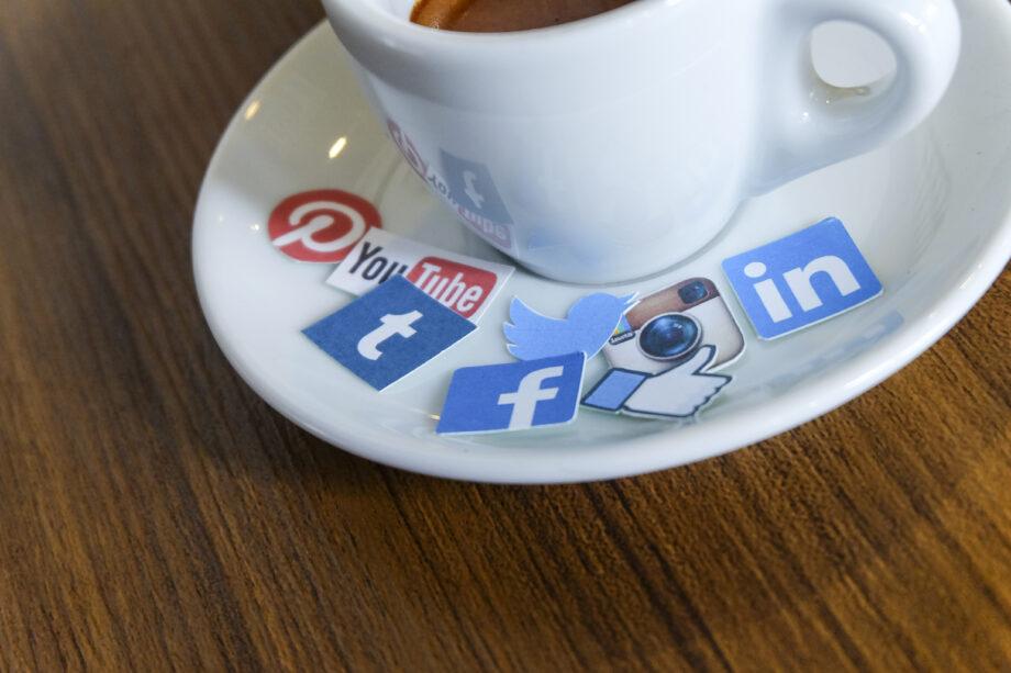 A white espresso cup on a saucer with social media logos like Pinterest, YouTube, Twitter, Instagram, LinkedIn, Facebook, and Tumblr cutouts scattered around it on a wooden table.