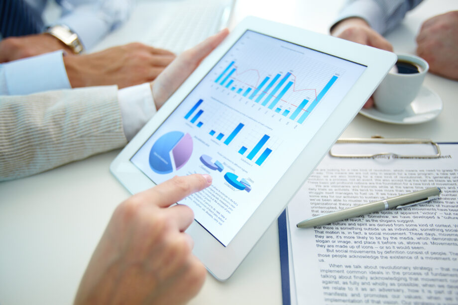 Hands hold a tablet displaying graphs and charts next to a document and pen on a table.