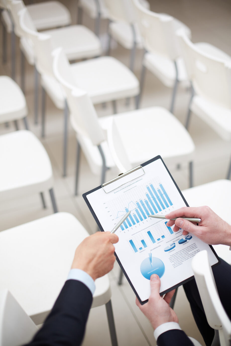 Two people in suits reviewing a clipboard with charts and graphs. White chairs are arranged in rows in the background.