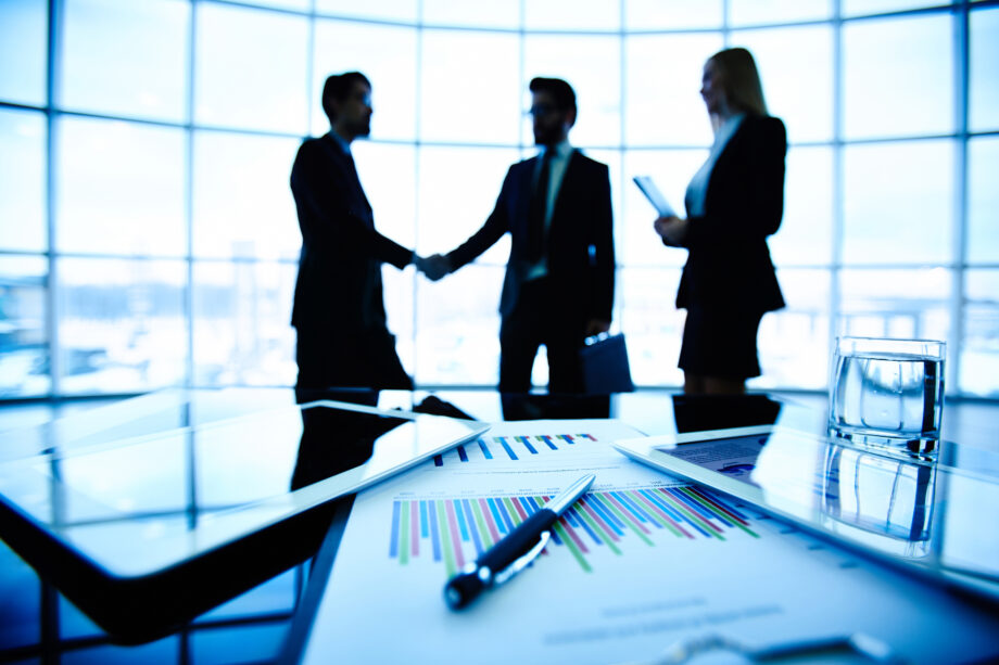 Silhouetted people in business attire in an office setting, with a handshake in the background. Documents, a pen, and tablets are on the table in the foreground.