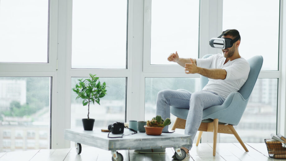 A man sits in a modern, brightly lit room wearing a VR headset, gesturing with his hands as if interacting with a virtual environment. Nearby is a table with plants, a tablet, and other items.