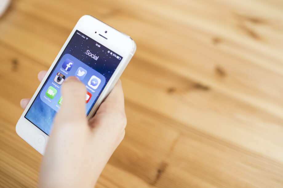 A hand holding a smartphone with visible social media app icons on the screen, including Facebook, Instagram, and Twitter, over a wooden table.