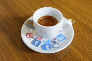 A cup of espresso on a saucer with social media logo stickers, placed on a wooden table.