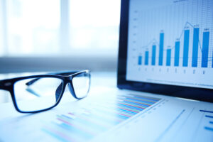 Close-up of eyeglasses resting on a financial report, with a laptop screen displaying a bar and line graph in the background.