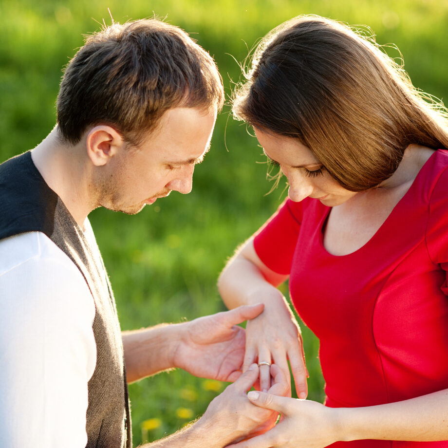 A man places a ring on a woman's finger in a grassy outdoor setting.