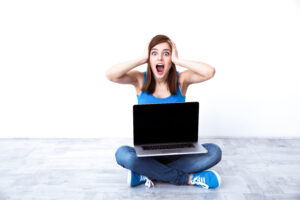 A person sitting cross-legged on the floor with a laptop, looking surprised, hands on their head, against a white background.