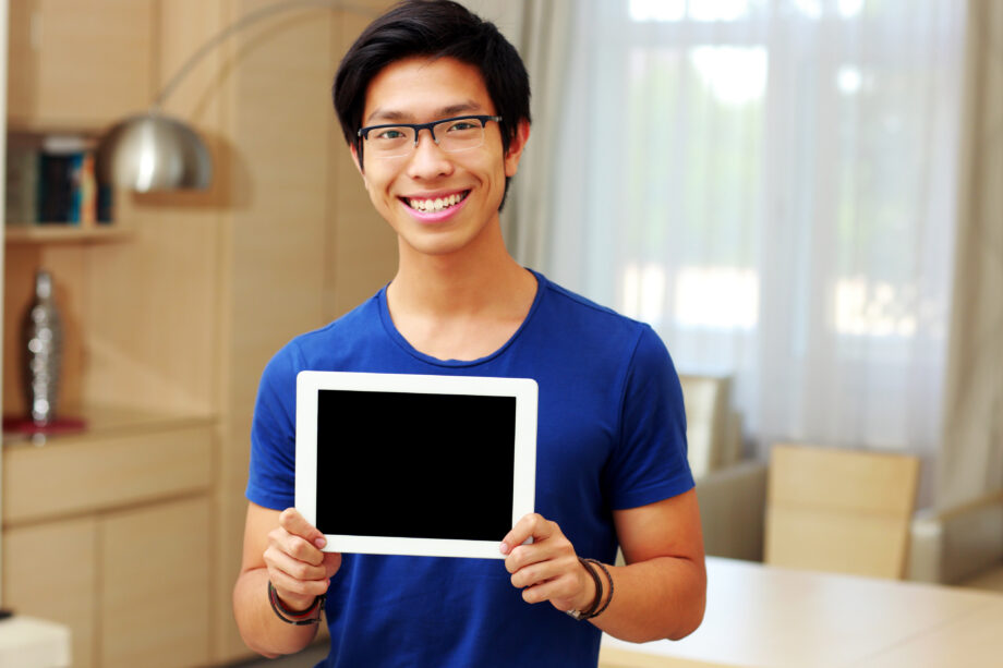 A person in a blue shirt smiling and holding a tablet with a blank screen indoors.