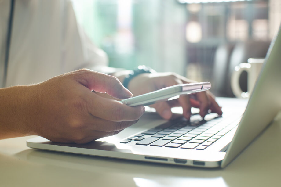 A person using a smartphone and typing on a laptop at a desk.