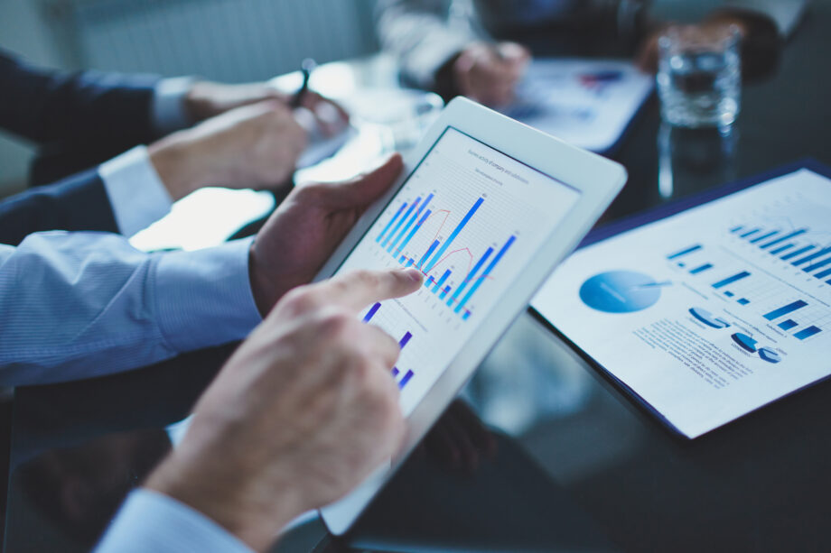 A person uses a tablet to view graphs and charts during a meeting, while another person takes notes.